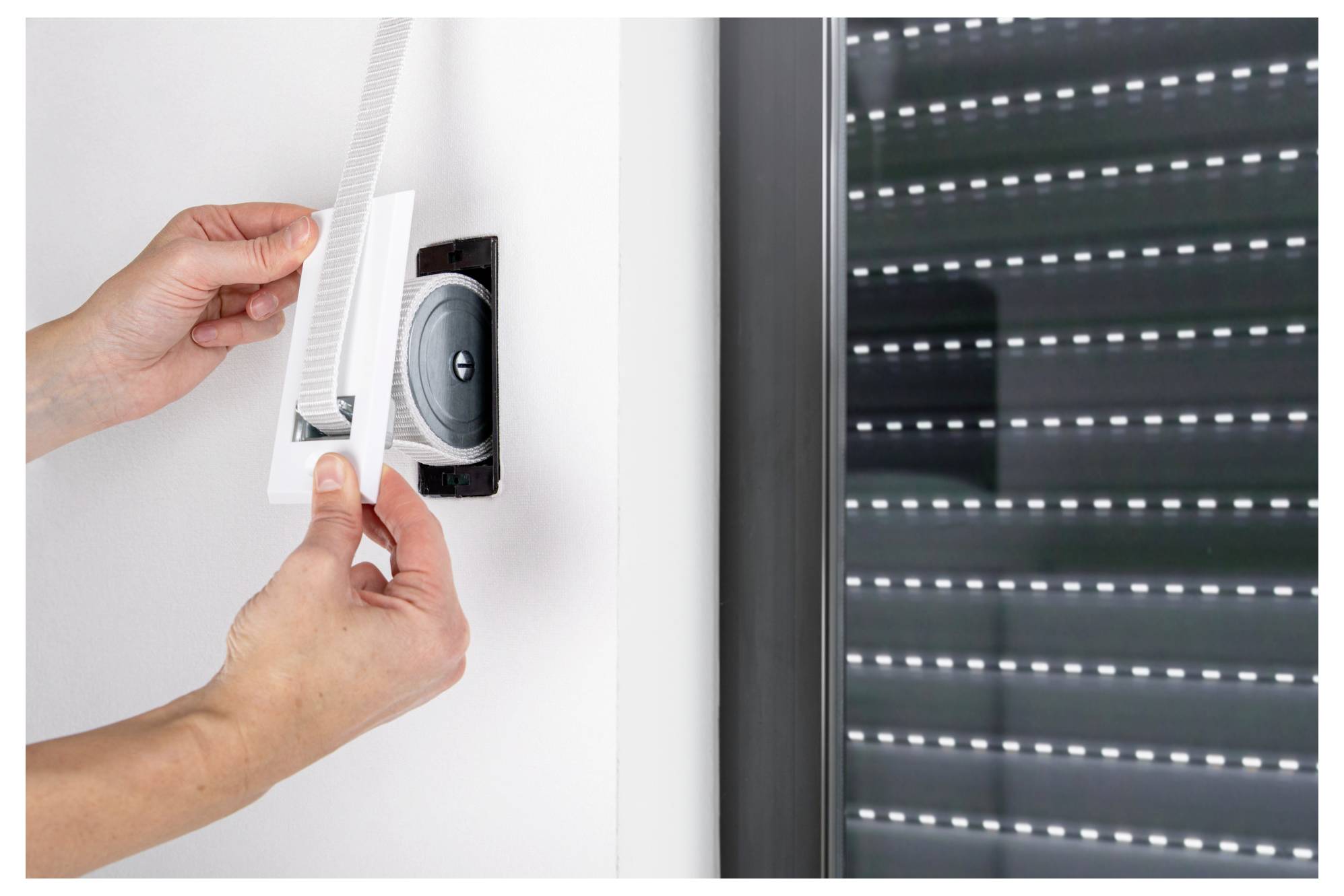 Hands opening a wall ventilation grille beside a window with a roller shutter, exposing the vent opening.<br>