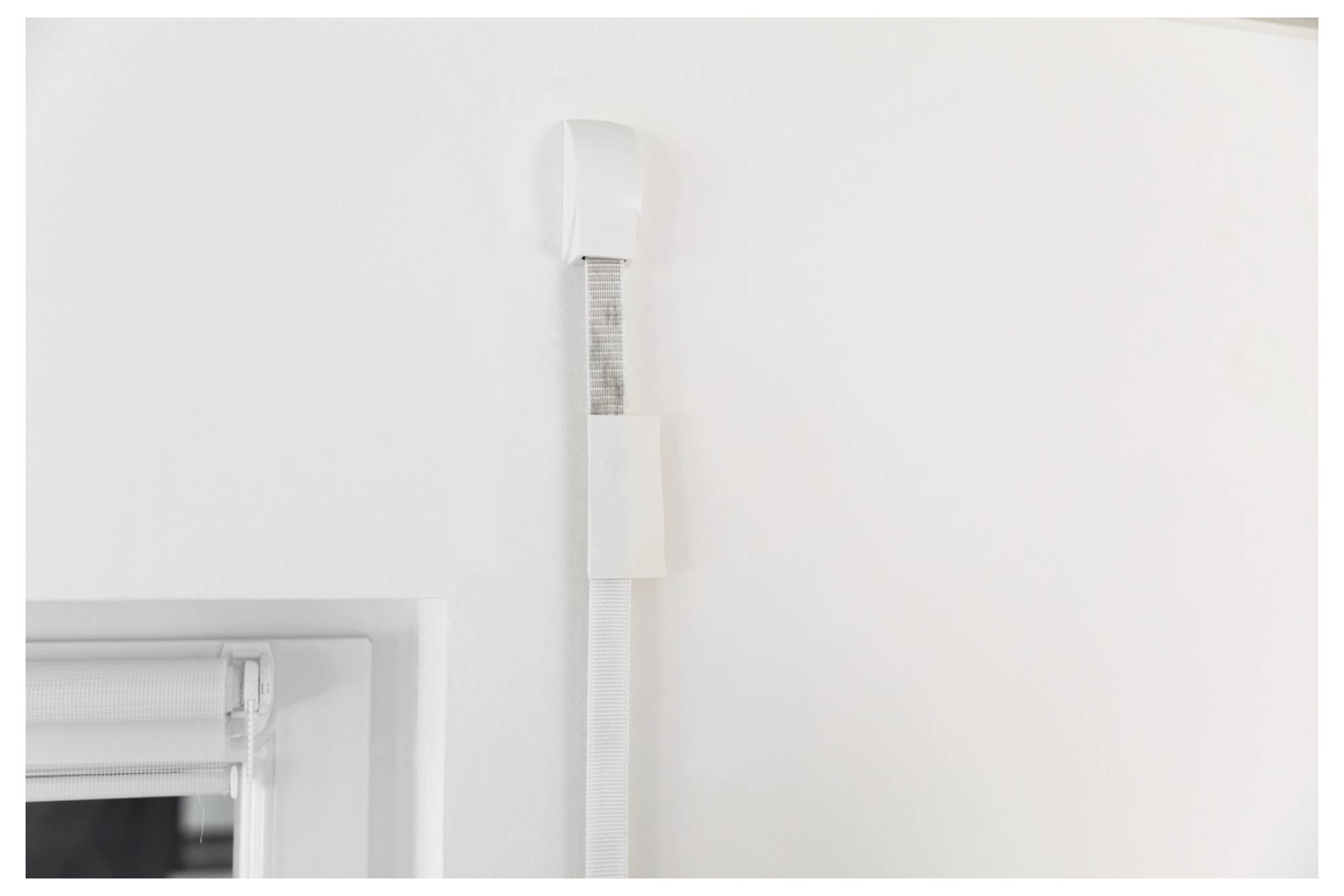 Close-up of a white wall-mounted cable channel or sensor casing beside a window frame on a plain white wall.