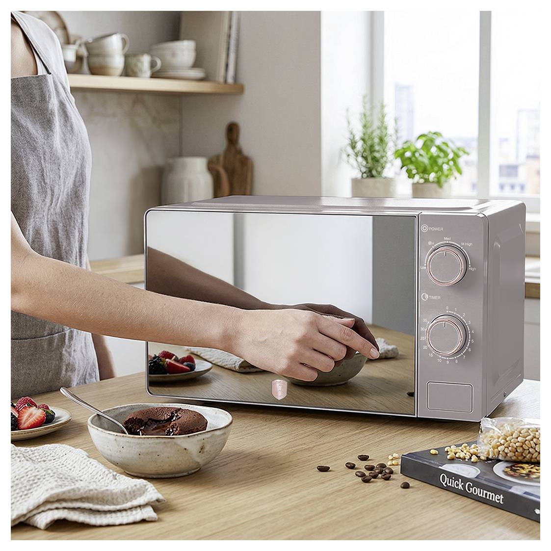 Person placing a bowl into a stainless steel countertop microwave in a bright kitchen, with food and ingredients on the table.<br>