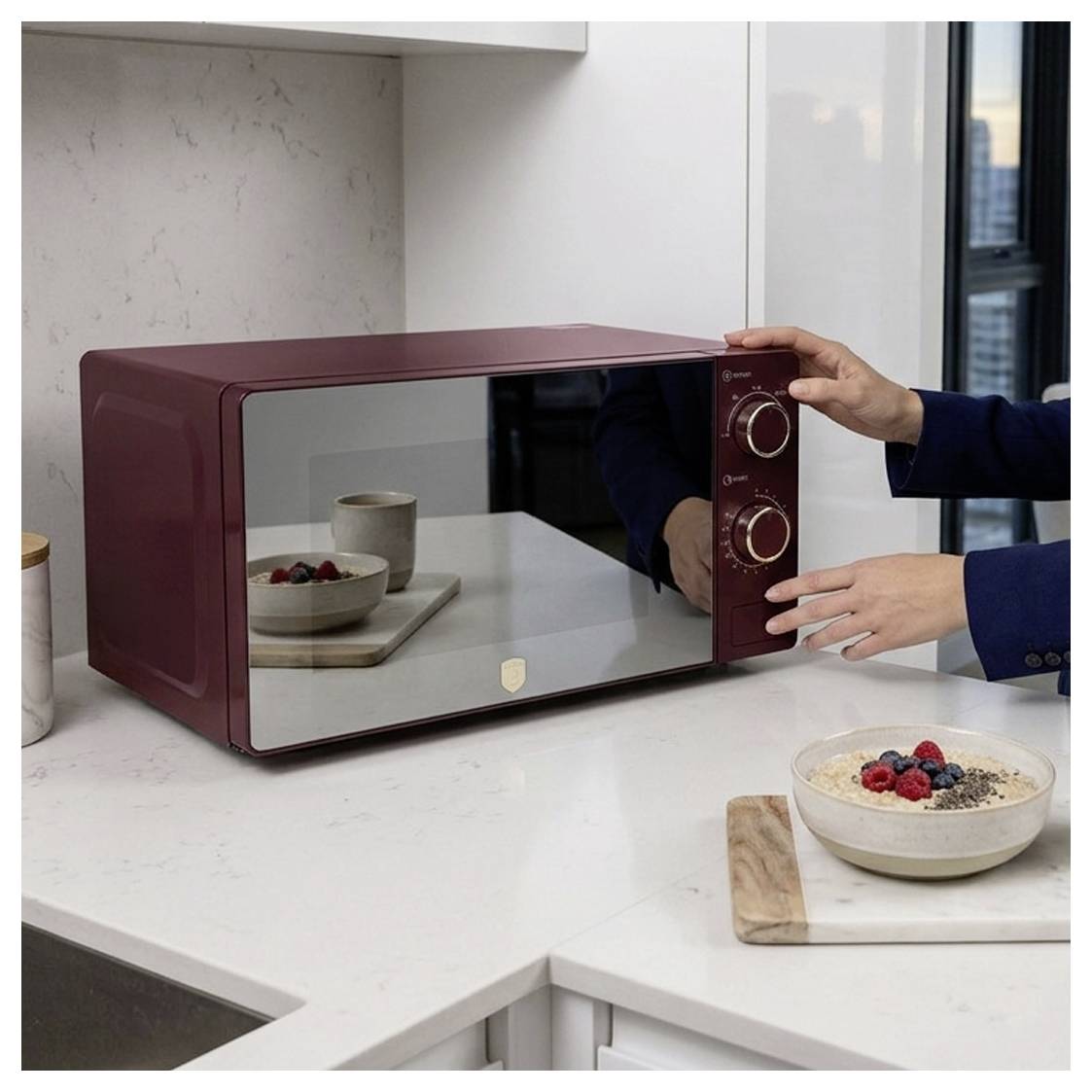 A maroon countertop microwave being adjusted by two hands in a modern kitchen, with bowls of oatmeal and berries nearby.