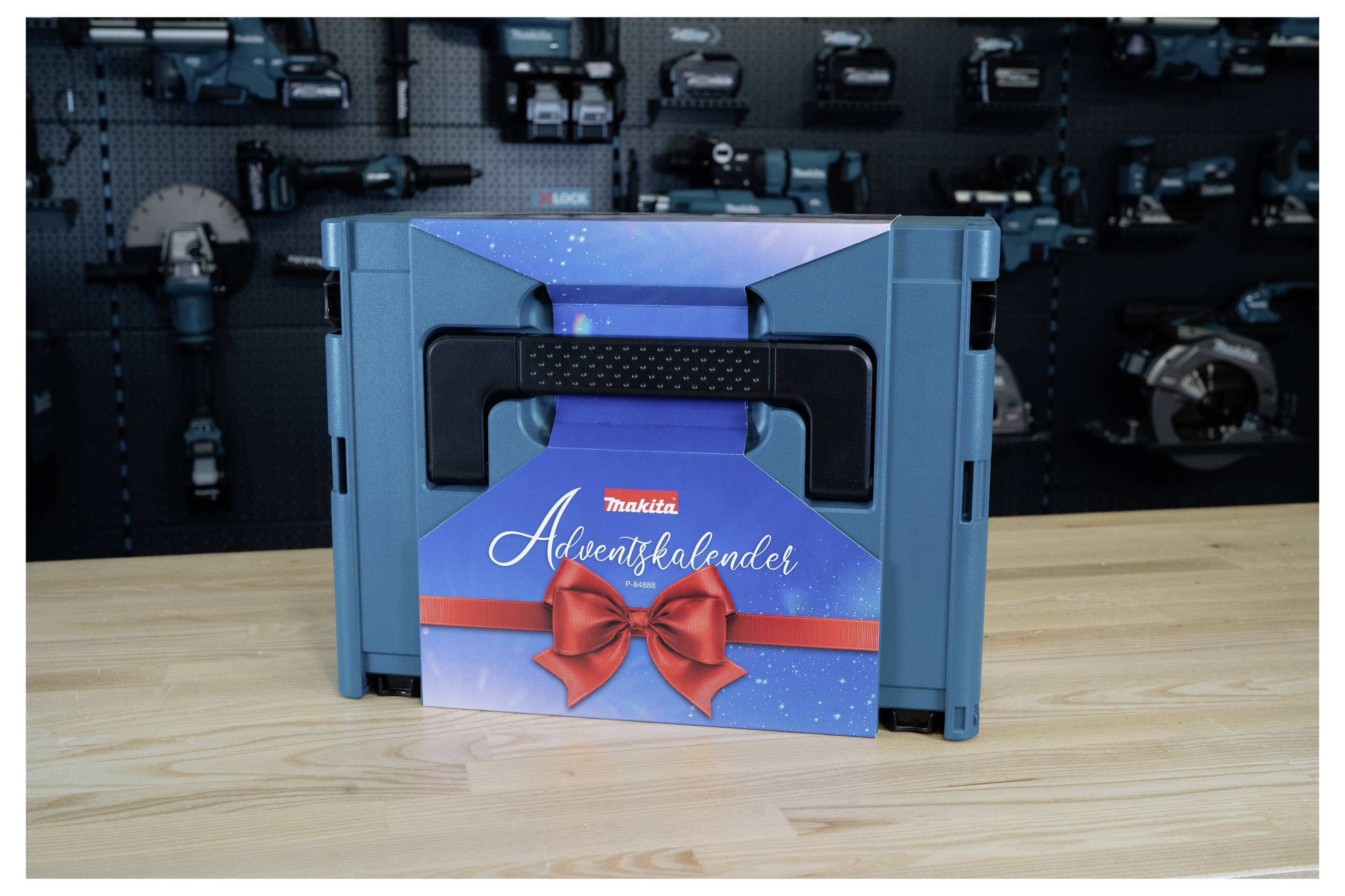 Blue Makita Adventskalender gift box with red ribbon and bow on a wooden table, in front of a tool display wall.<br>