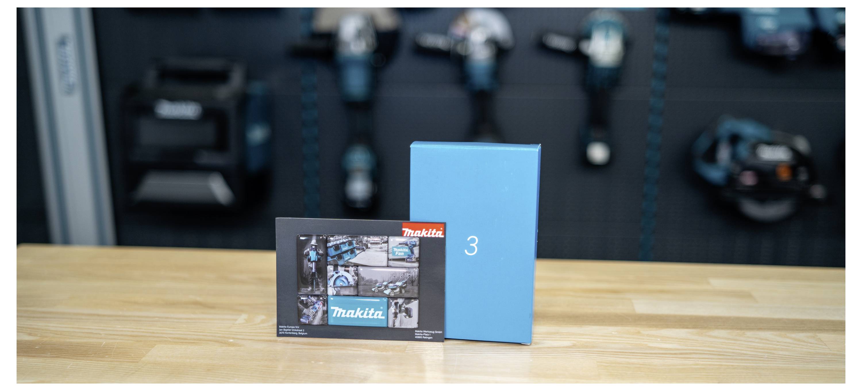 Makita product box and a blue box labeled '3' on a wooden table, with blurred power tools in the background.