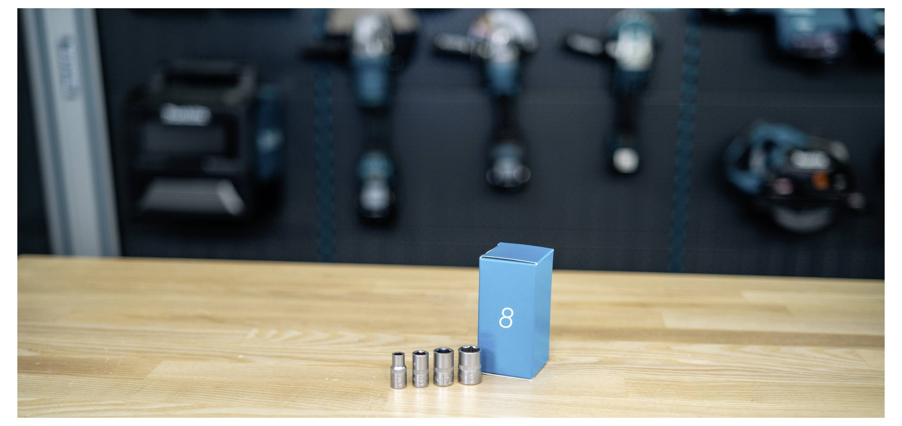 Blue box labeled '8' beside four small metal battery cells on a wooden table, with blurred tools in the background.
