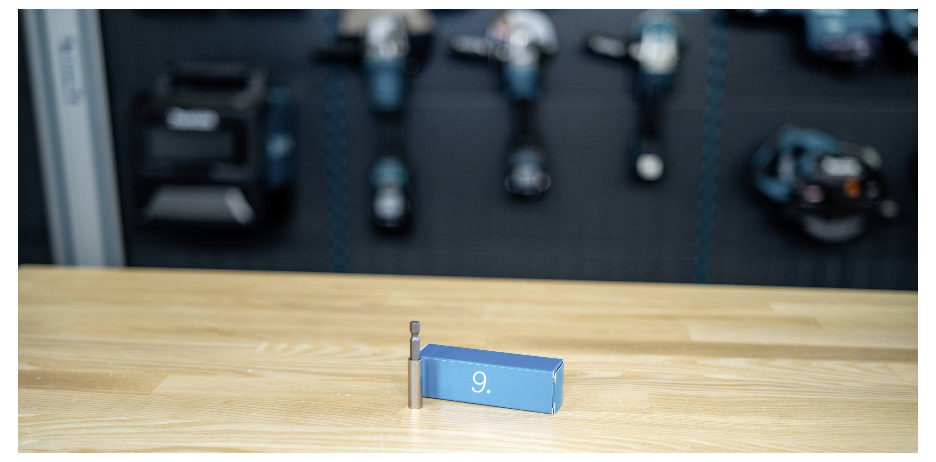 Small blue block labeled '9' with a peg on a wooden surface, against a blurred background of stacked dumbbells and gym equipment.