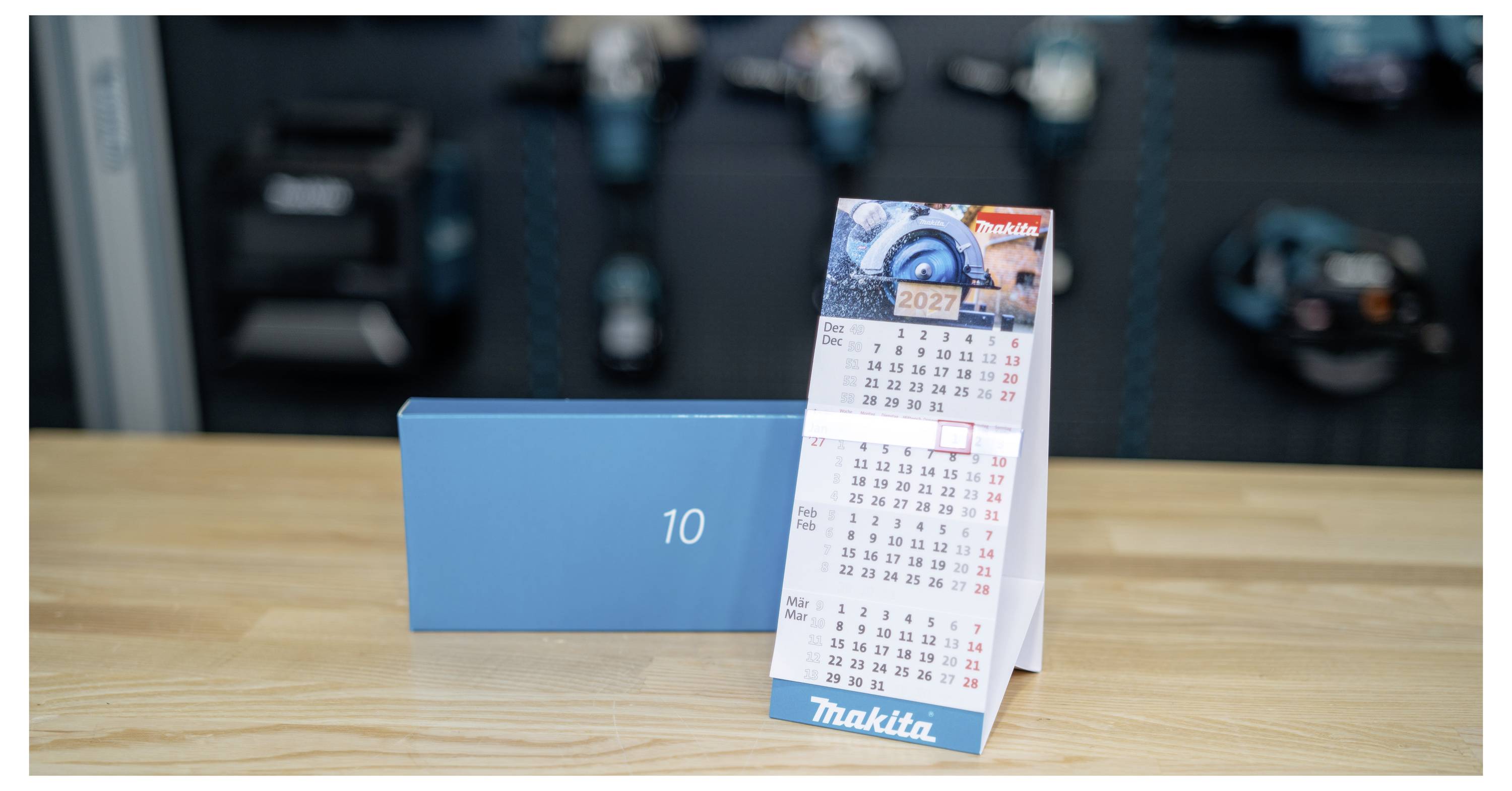 A Makita desk calendar stands on a wooden table beside a blue box labeled '10', with blurred tool-like objects in the background.