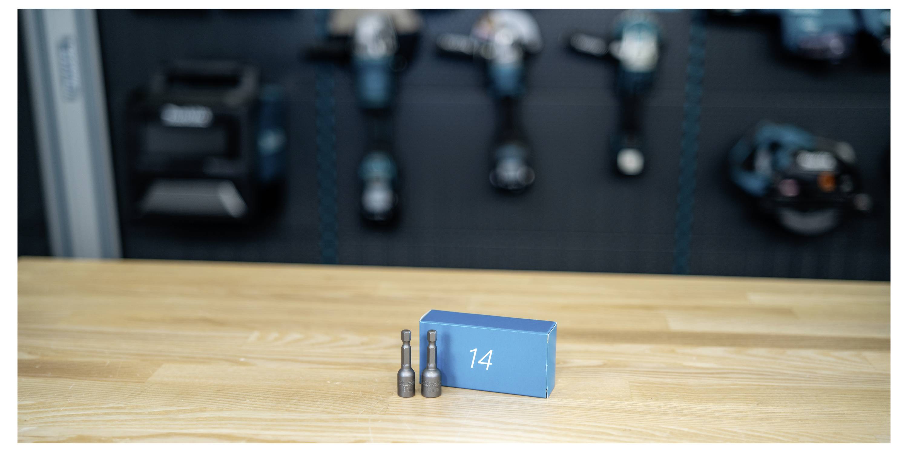 Numbered blue block '14' with two metal bolts on a wooden table, with blurred industrial machinery in the background.