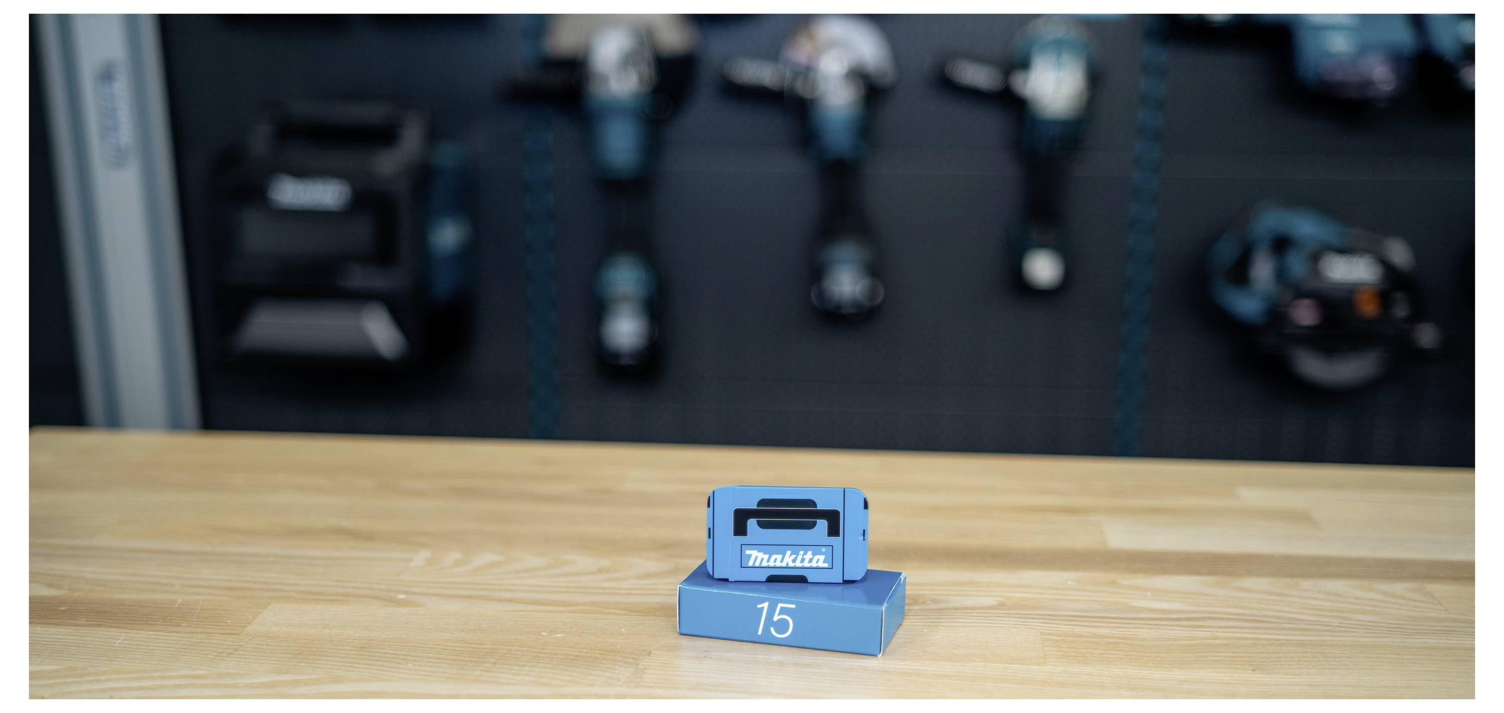 A blue Makita battery charger sits on a wooden workbench, with blurred power tools and equipment in the background.