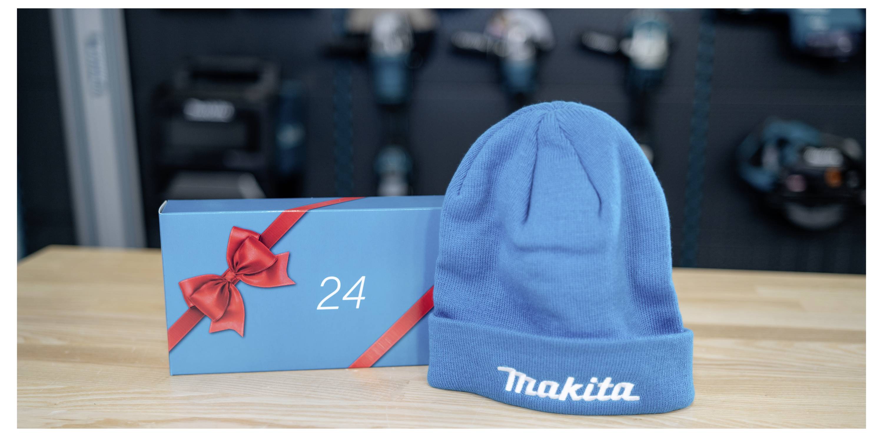 Blue Makita beanie hat beside a blue gift box with a red ribbon and the number 24 on a wooden table, with blurred machinery in the background