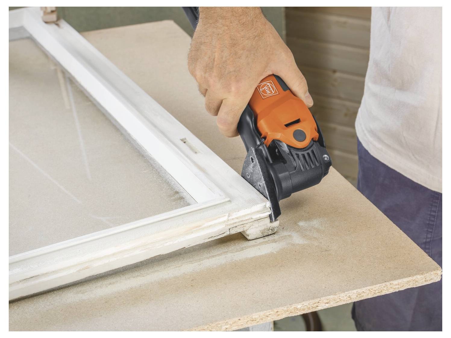 Person uses an orange power tool to cut the corner of a white window frame on a workbench.