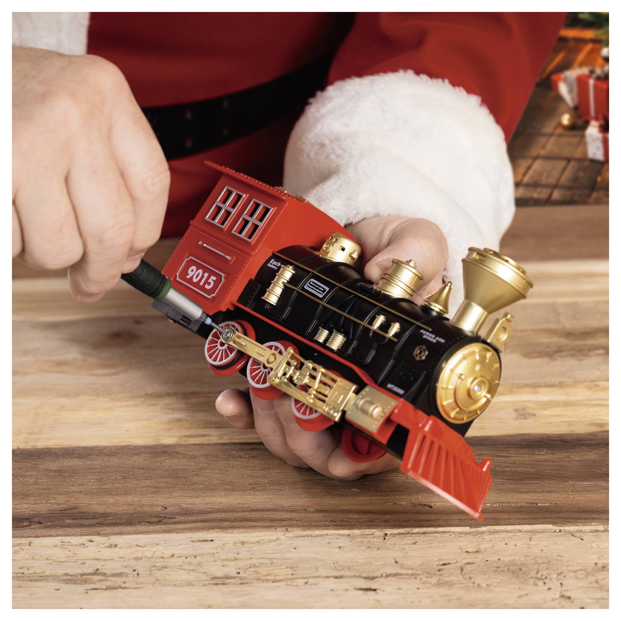 Hands hold a red-and-black toy steam locomotive on a wooden table, with a small '9015' label on the engine.<br>
