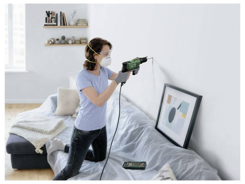 Woman wearing a face mask drills into a bedroom wall while kneeling beside a bed, with a framed picture leaning nearby.