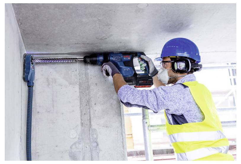Construction worker in hard hat, safety vest, and mask drilling into a concrete wall with a power drill.