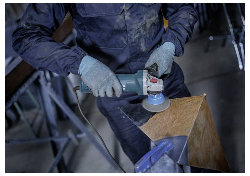 Gloved worker uses an electric angle grinder to sand a metal piece clamped in a workshop.
