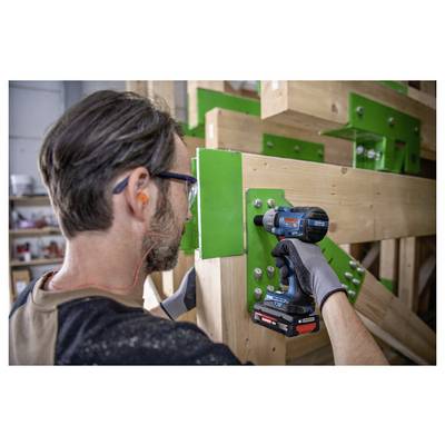 Worker using a cordless drill to fasten a green metal connector onto a wooden beam in a workshop.