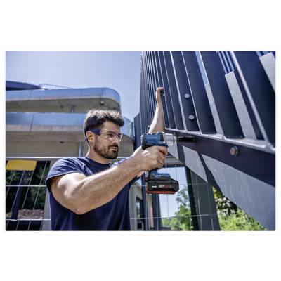 A man using a cordless drill to work on a large metal structure outside a modern building.<br>