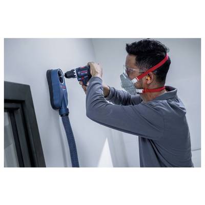 Technician wearing safety glasses and mask drills into a wall beside a mounted blue vacuum dust collector.<br>
