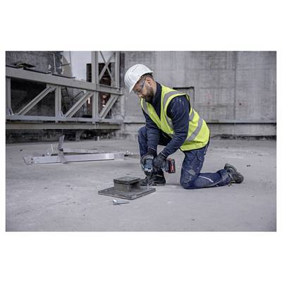 Construction worker in a hard hat and safety vest kneels on concrete, using a power drill to fasten a metal base plate at an industrial site.
