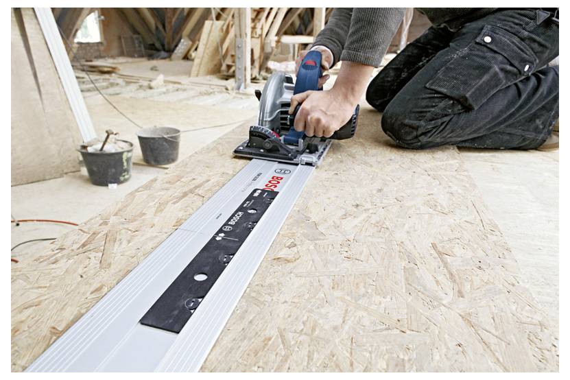 Carpenter kneeling on plywood cuts a board with a circular saw guided by a long straight edge in a construction site.<br>