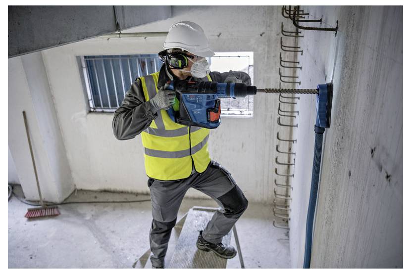 Construction worker in hard hat and safety vest drilling into a concrete wall inside an unfinished building.