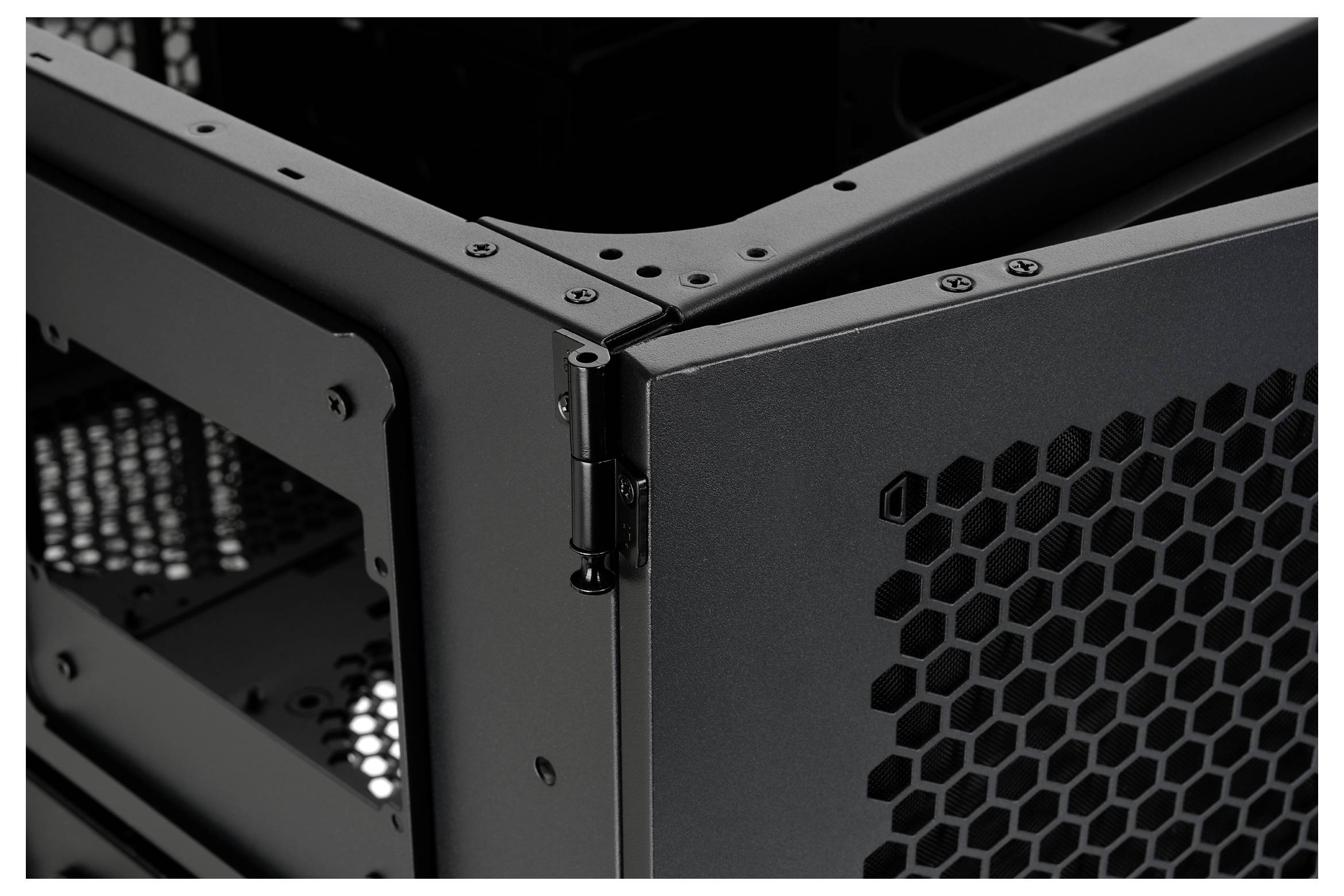 Close-up of a black computer case corner with a front hexagonal mesh panel and visible hinge, showing the chassis frame and ventilation design.<br>