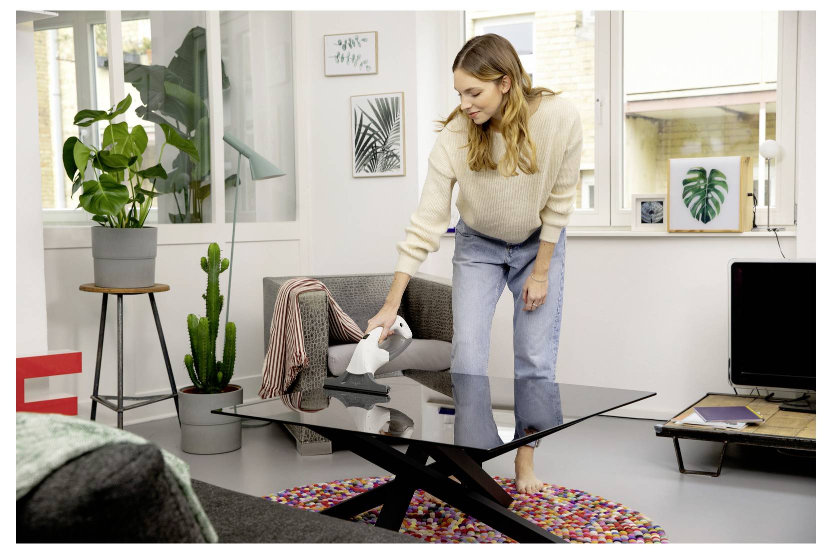 Woman vacuuming a glass coffee table in a bright living room with plants and wall art.