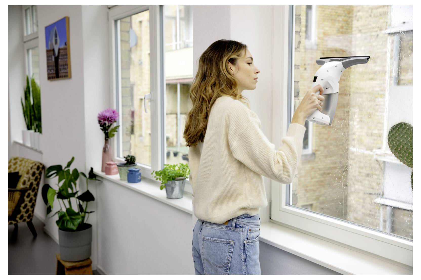 Woman cleaning a window with a handheld squeegee in a bright room with potted plants on the sill.<br>