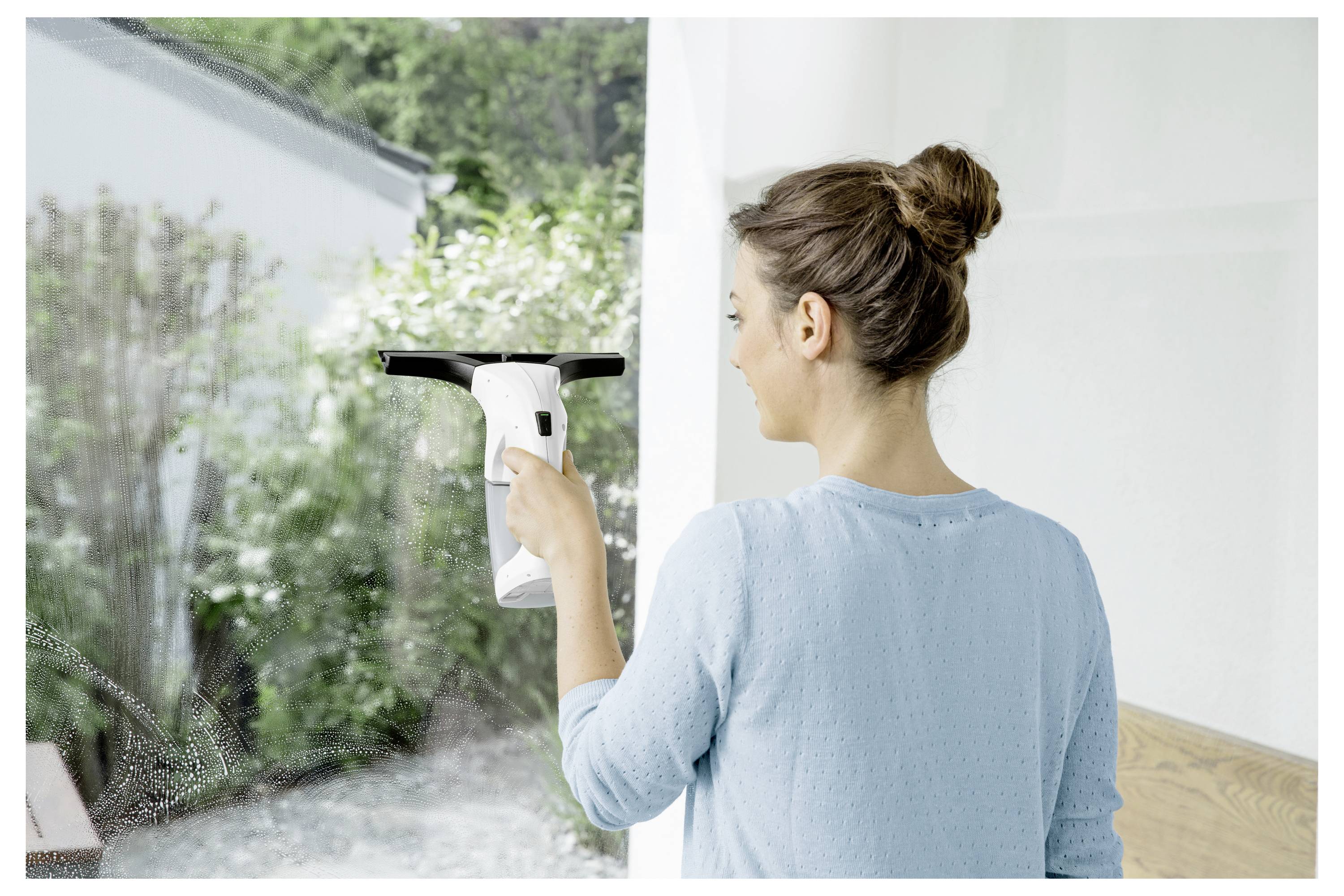 Woman cleaning a large window with a handheld spray cleaner, seen from behind in a bright room.<br>
