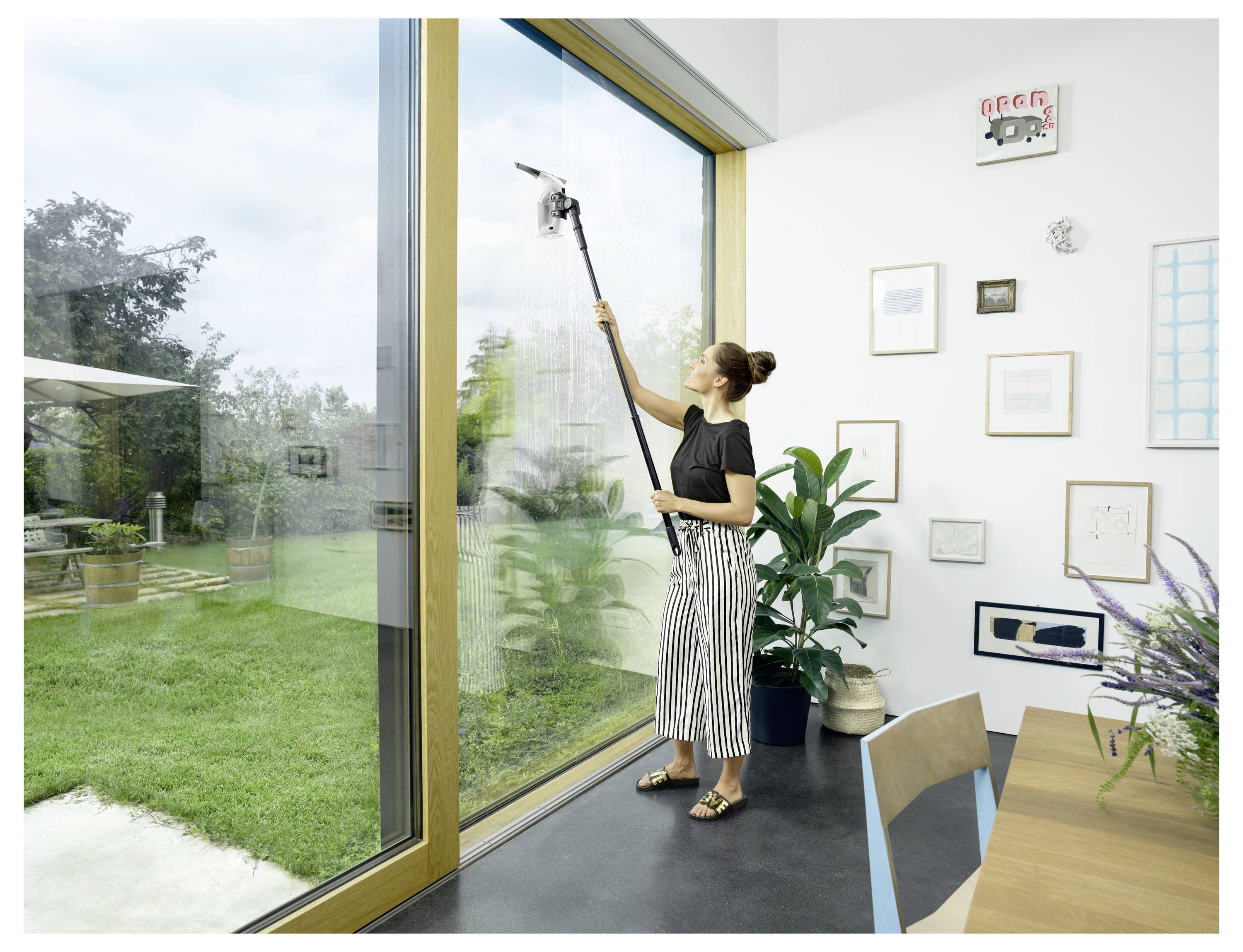 Woman using a long-handled window squeegee to clean a large floor-to-ceiling glass door in a bright room overlooking a garden.