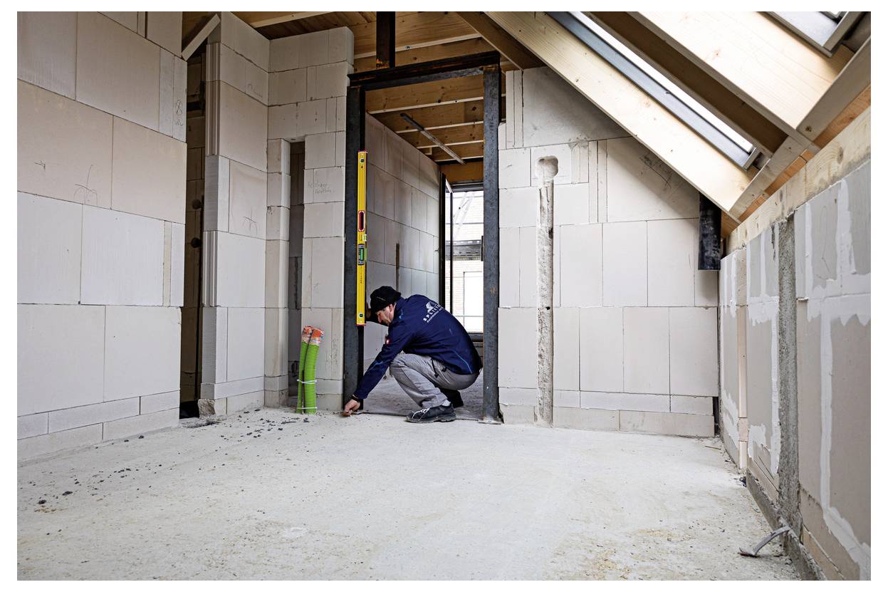 Worker measures an unfinished room with a yellow level during interior construction, showing exposed walls, beams, and floor under renovation.