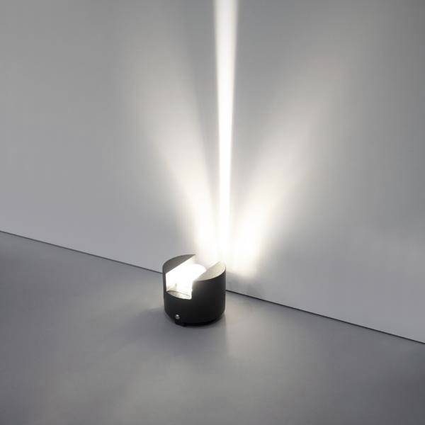 Small black wall light on a gray floor shining bright white beams upward onto a plain wall.<br>