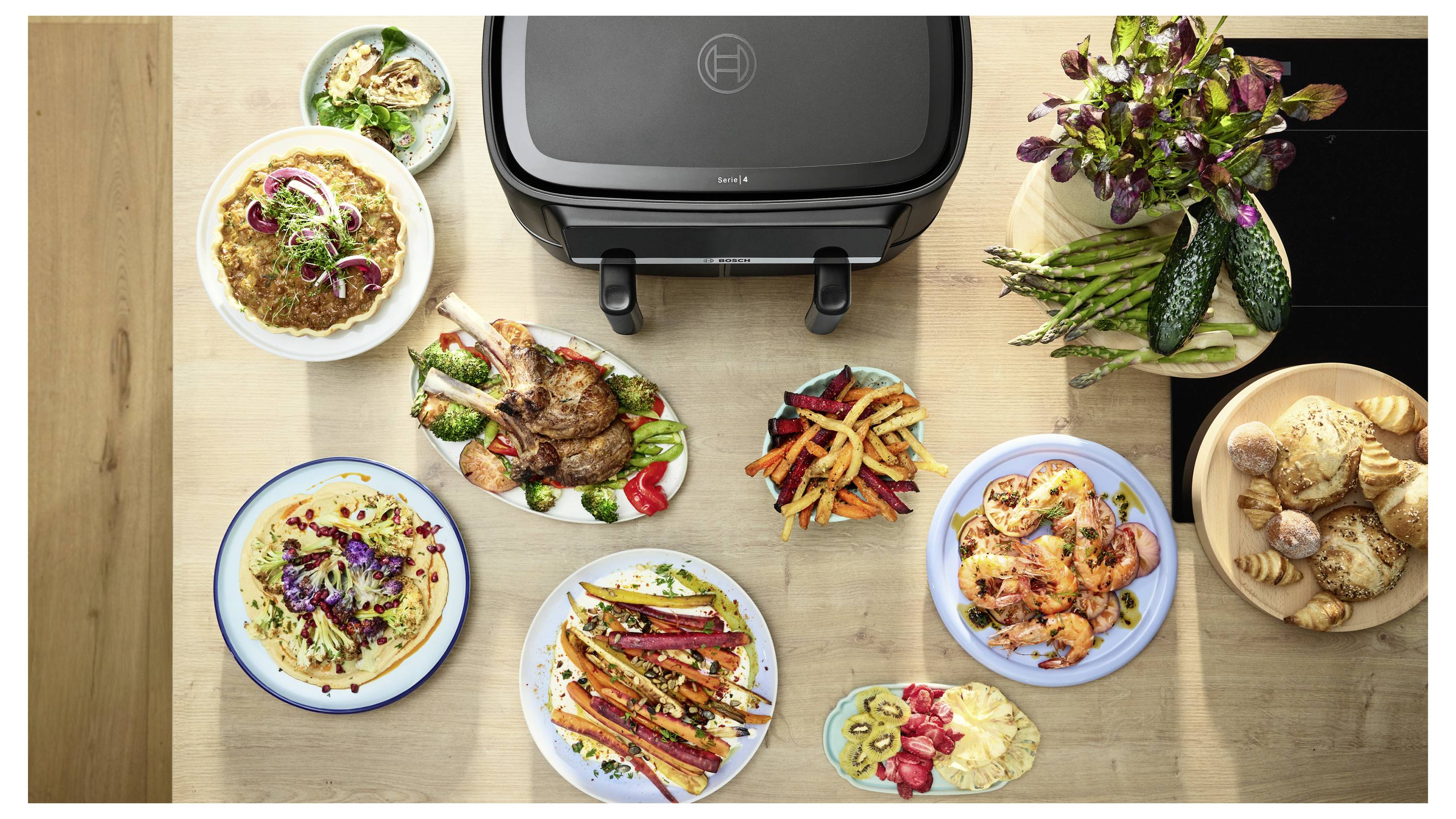 Top-down view of an electric grill surrounded by plated grilled dishes, roasted vegetables, and sides on a wooden table.