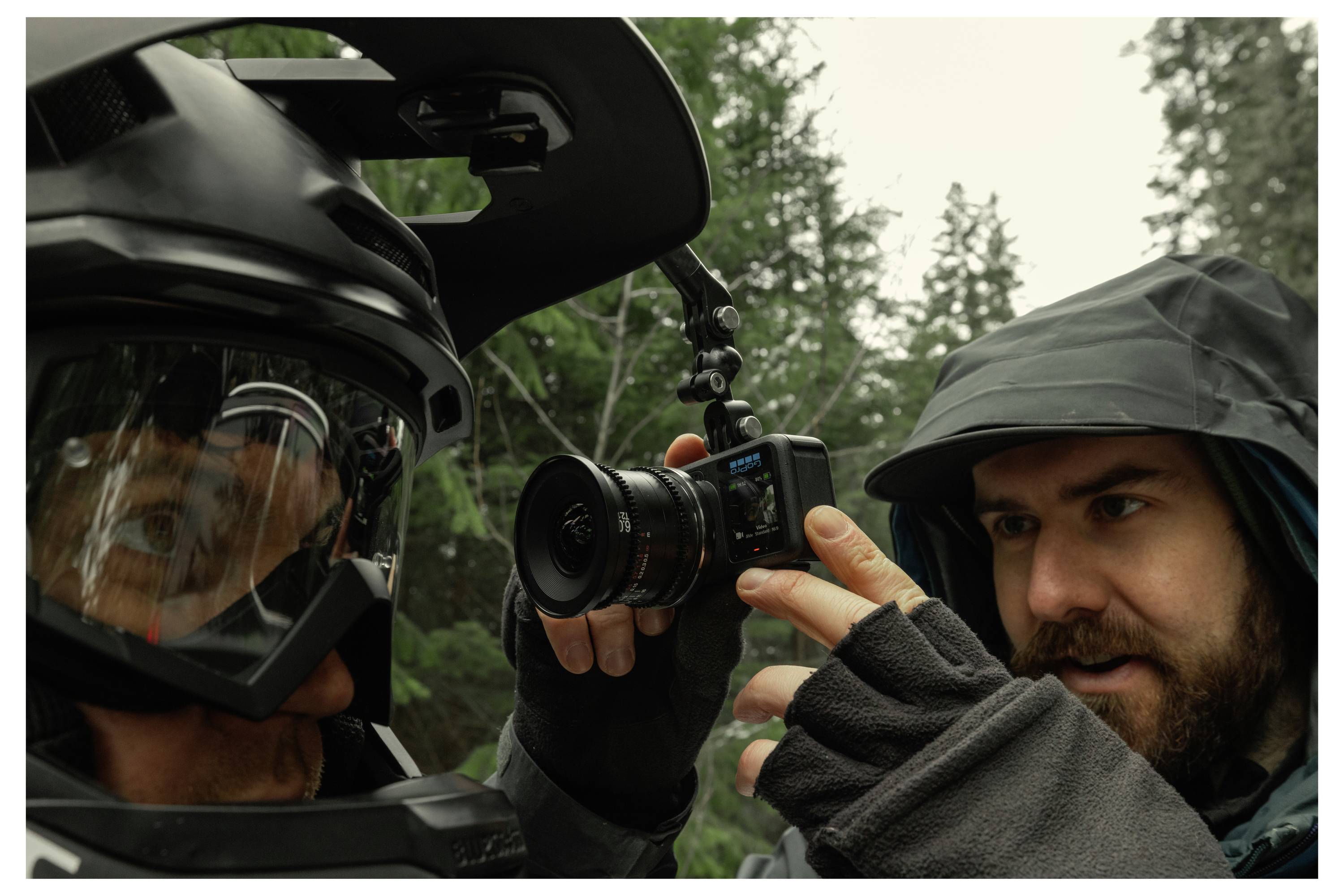 Close-up of a man in a hood filming a helmeted person with a camera in a forest, likely documenting a ride or outdoor activity.