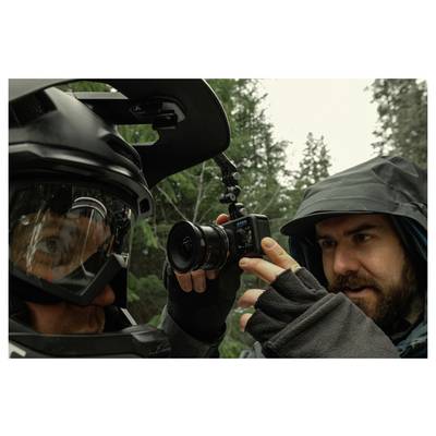 Close-up of a man in a hood filming a helmeted person with a camera in a forest, likely documenting a ride or outdoor activity.