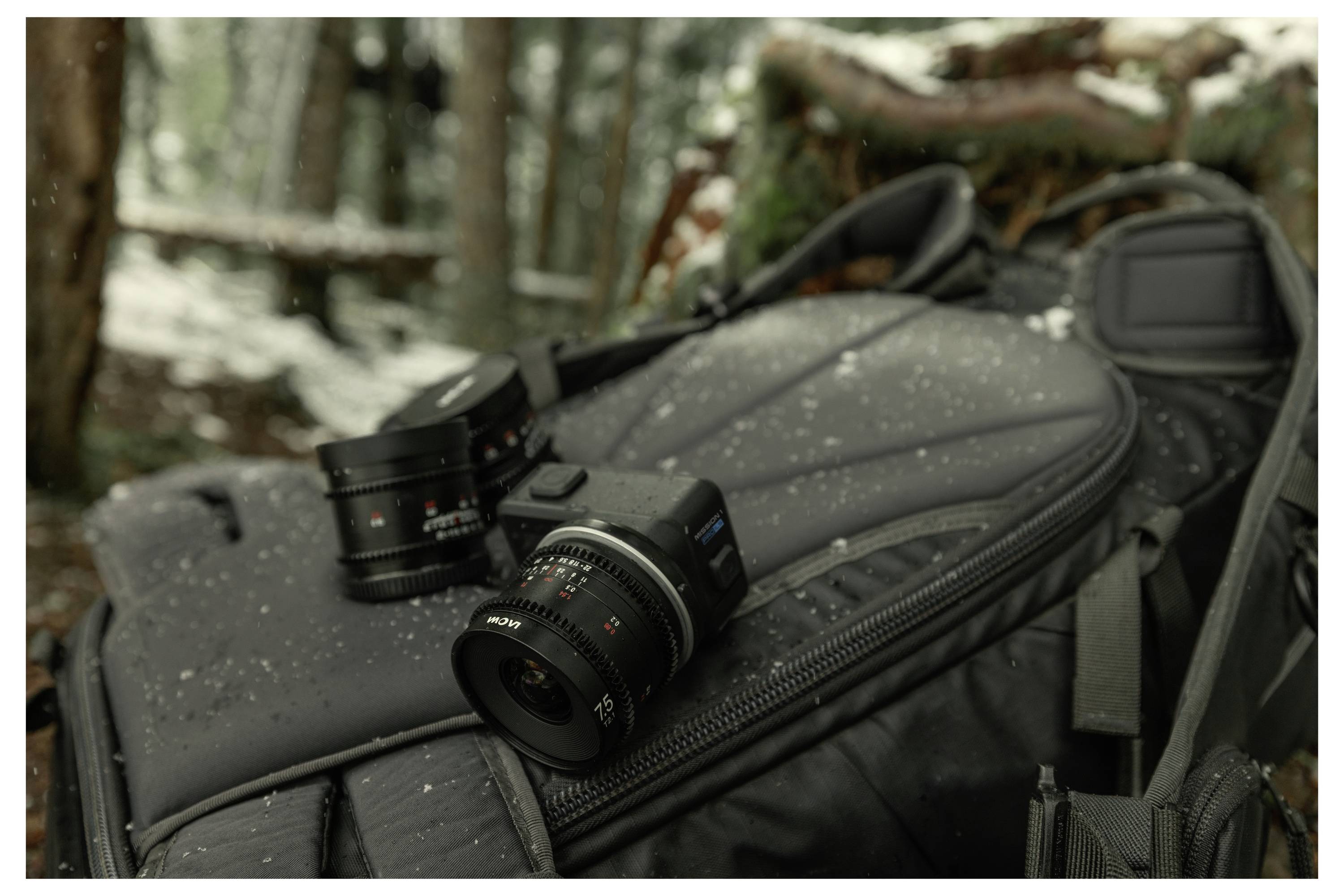 Camera lenses resting on a black backpack dusted with snow in a forest setting.