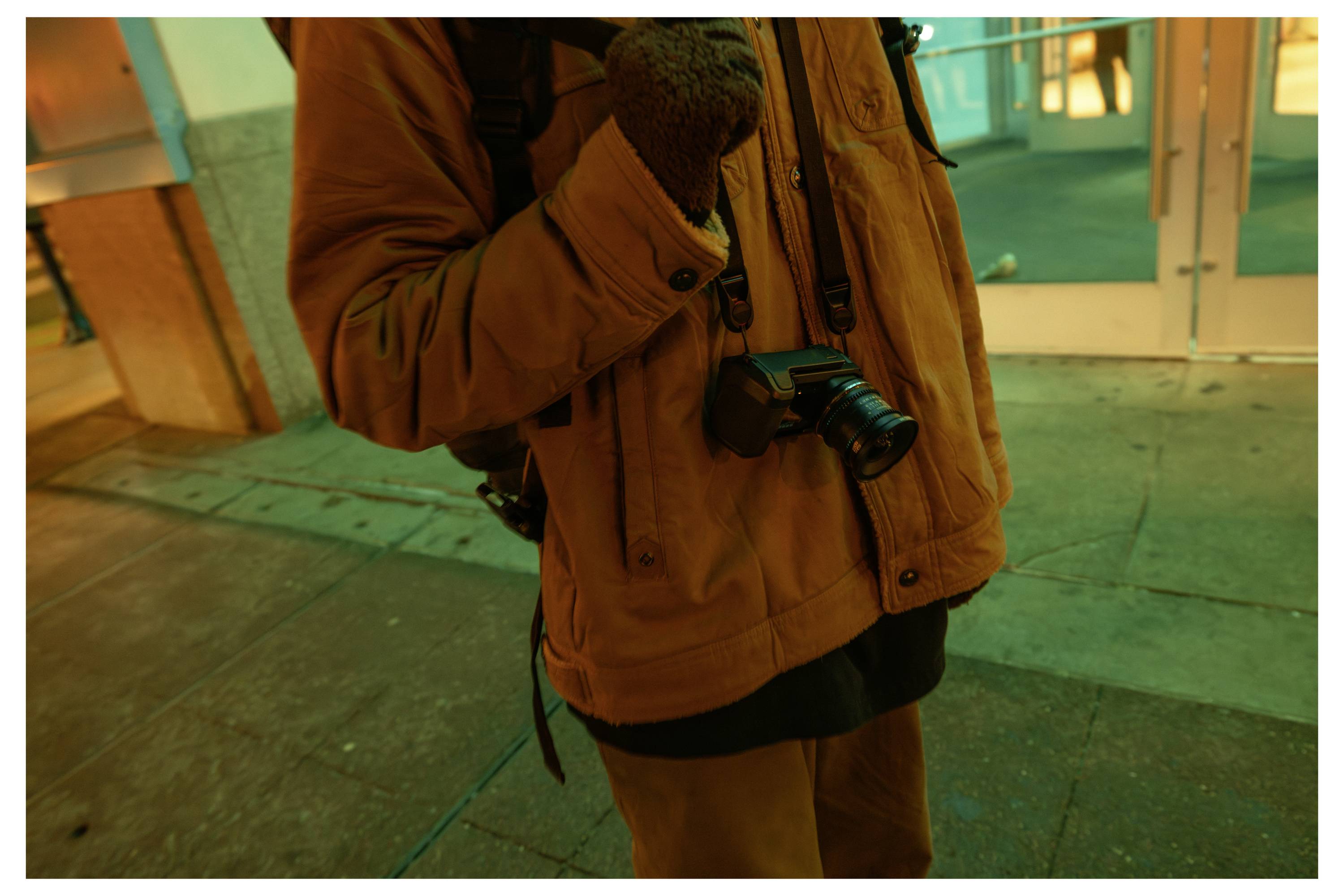 Person in a brown jacket and gloves holding a camera at night outside a building, with another camera hanging from their neck.