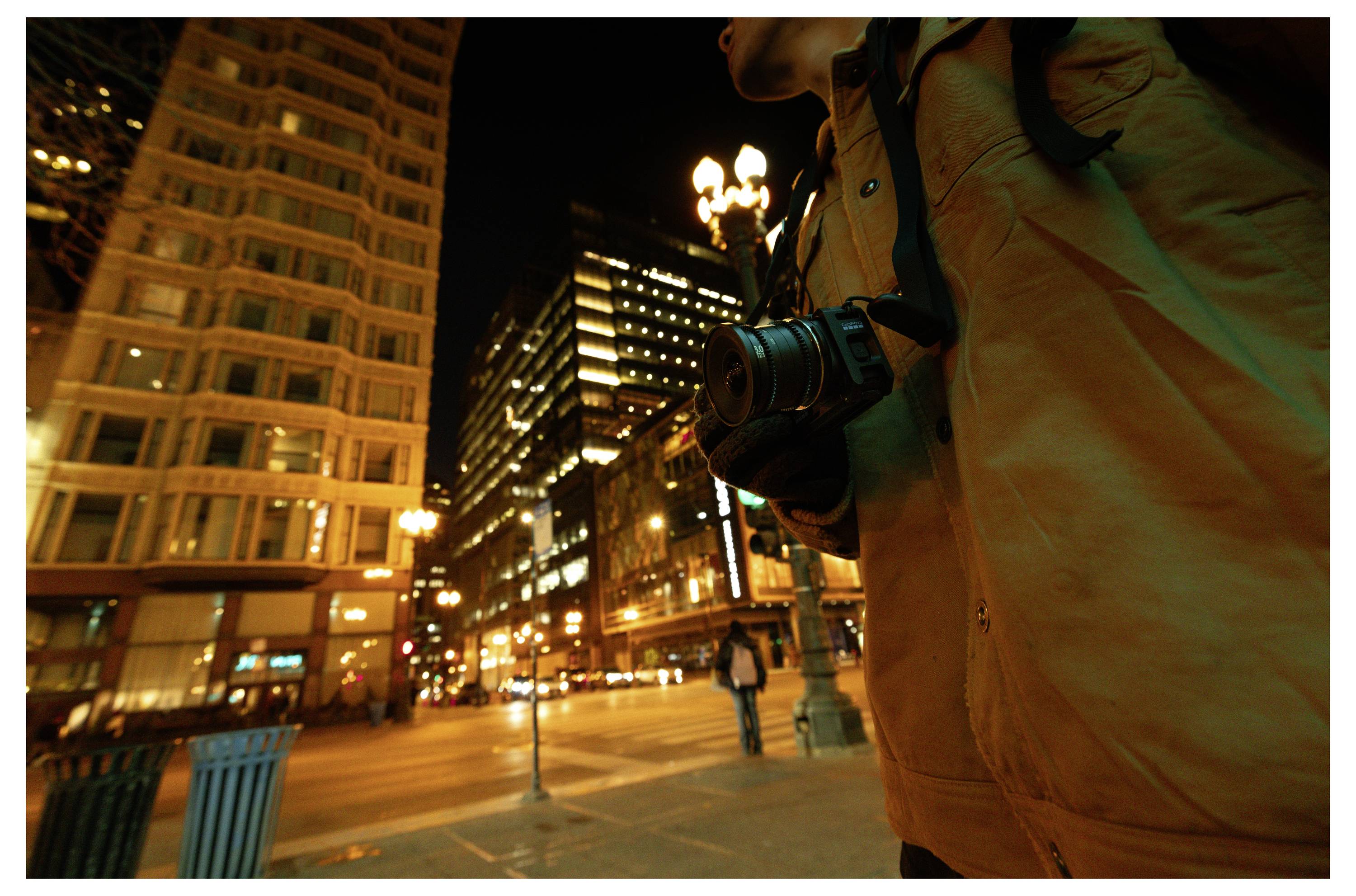 Person in an orange coat holding a camera on a nighttime city street with lit buildings and streetlights.<br>