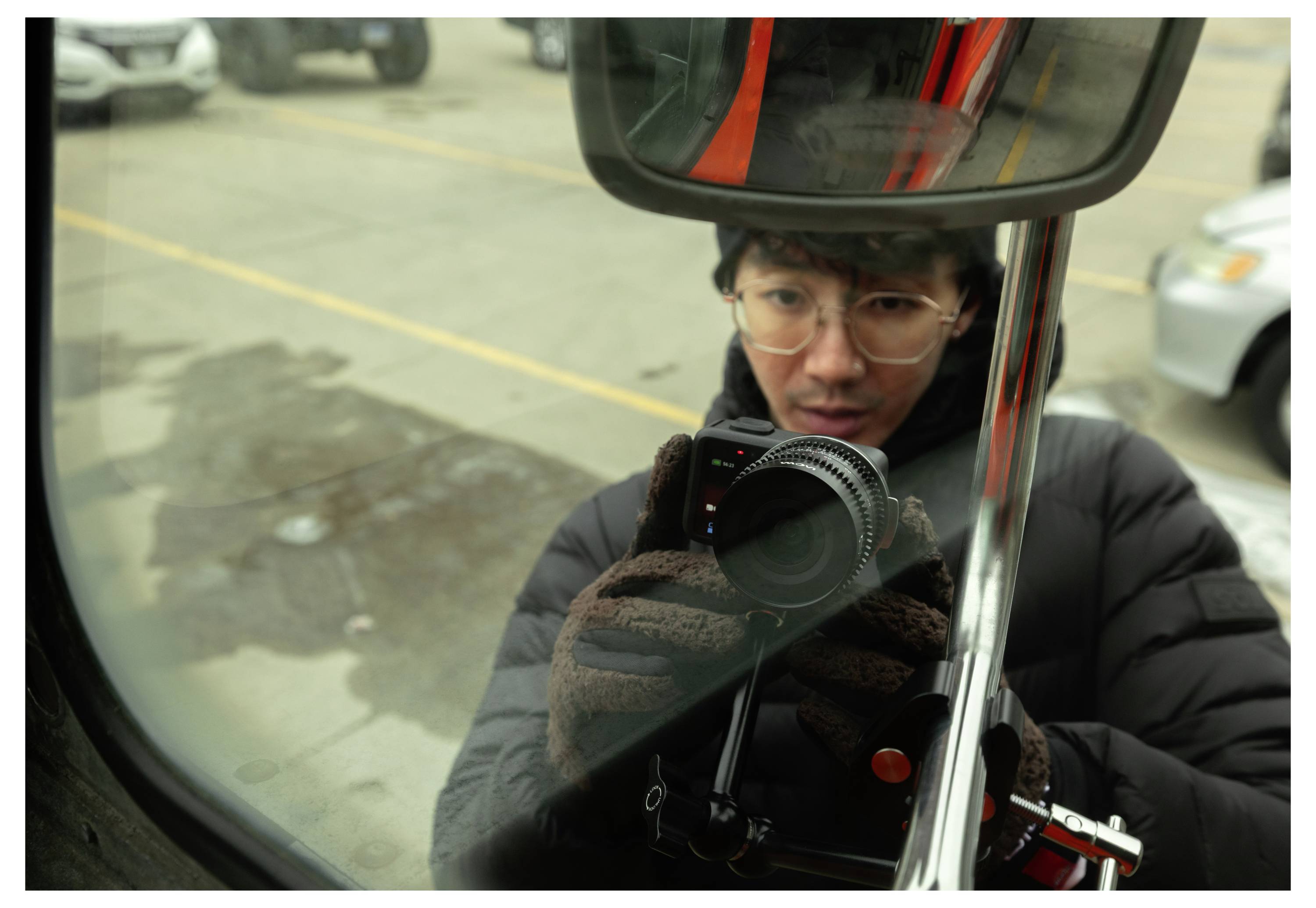 A photographer in glasses and a black jacket holds a camera, reflected in a vehicle mirror on a city street.<br>