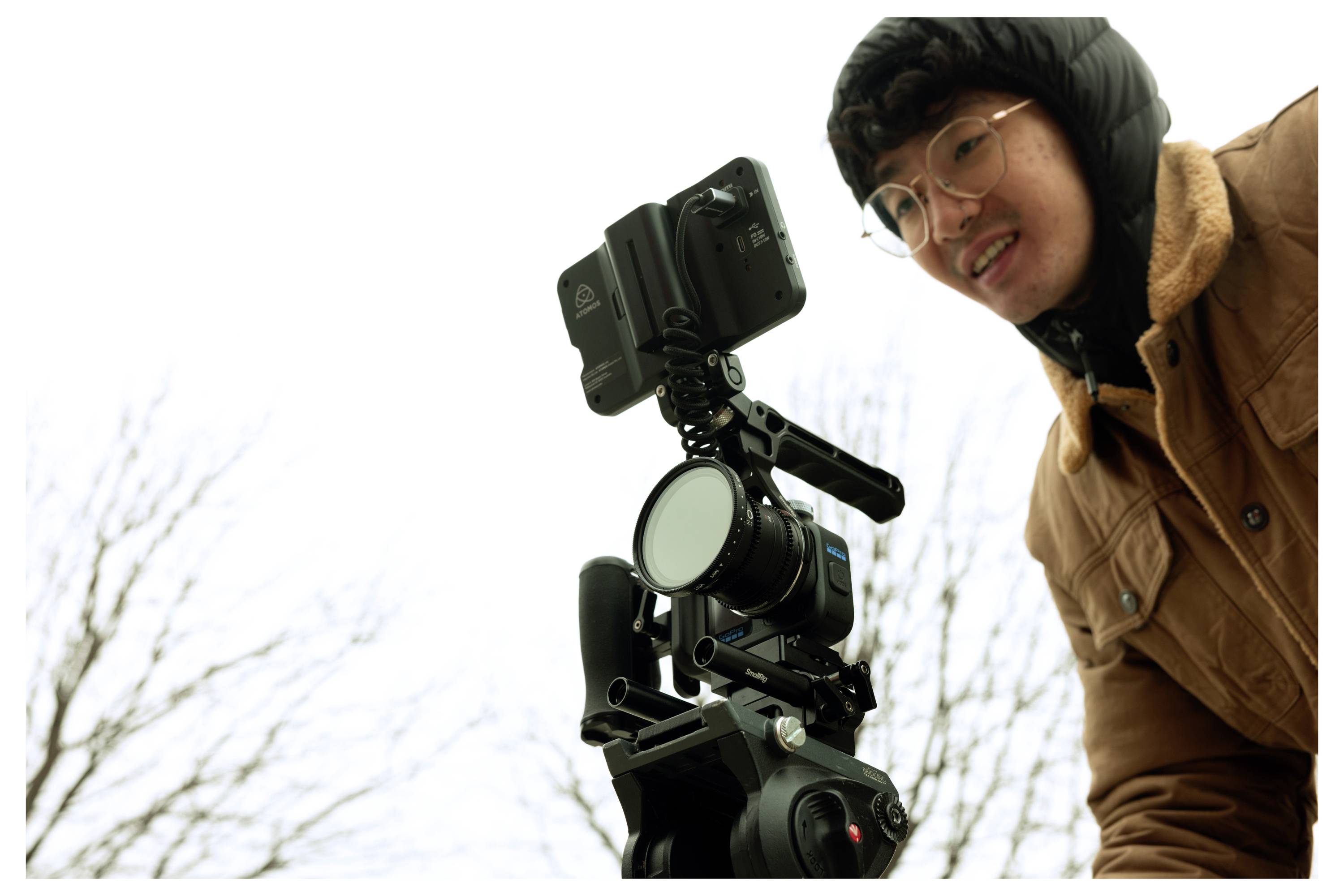 Man adjusting a camera mounted on a tripod outdoors on a cloudy day.