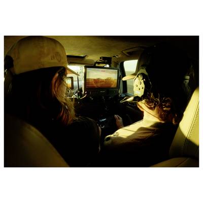 Two people sit in a small aircraft cockpit at sunset, with a front display showing a runway or landing view.<br>