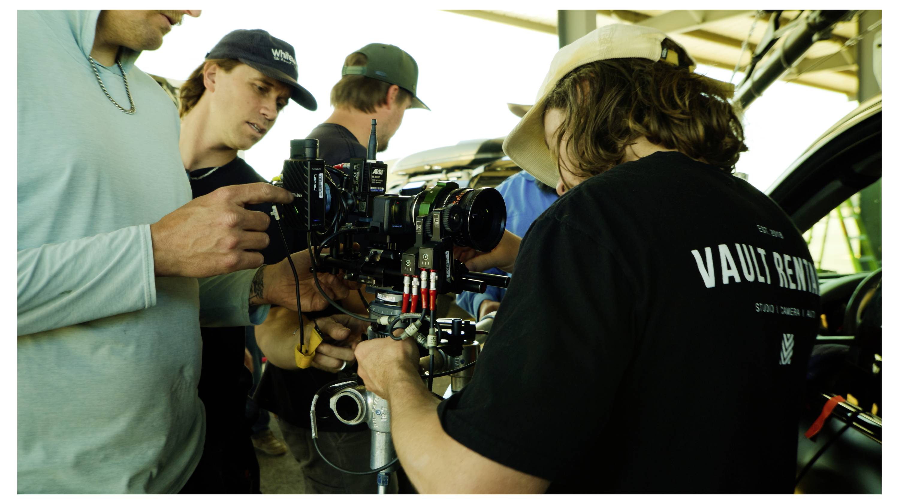 Film crew members adjust a camera on a car set, with one person in a 'Vault' shirt operating the rig.