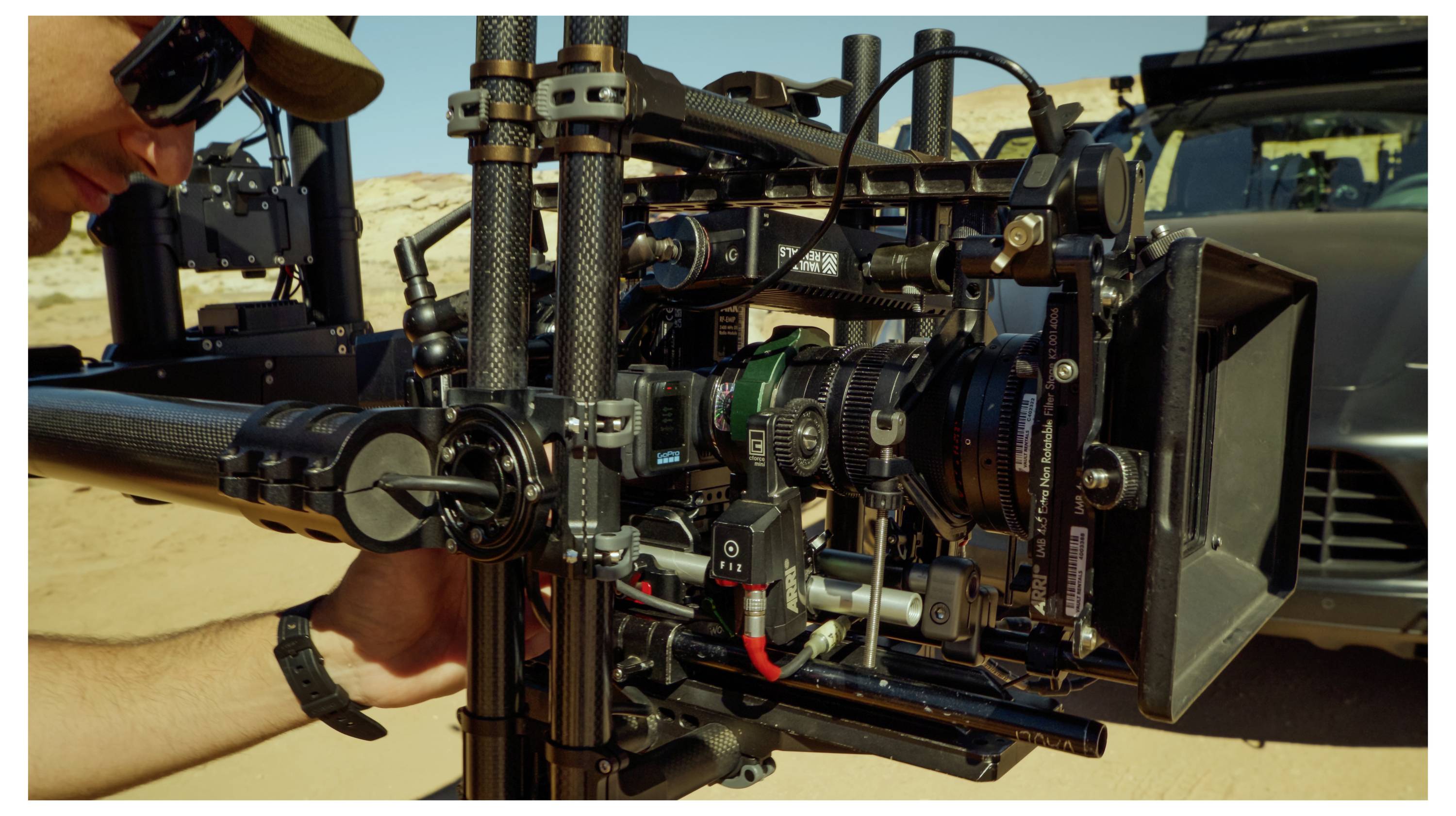 Person adjusting a professional camera rig with lens, follow-focus, and matte box outdoors beside a vehicle.