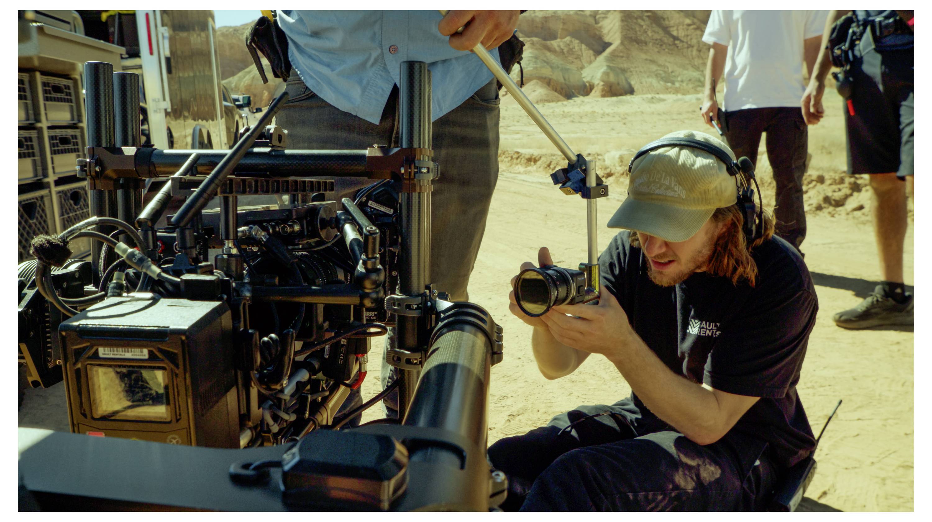 Camera operator kneels beside a film camera on set, adjusting the lens in a sandy outdoor scene while crew members stand nearby.