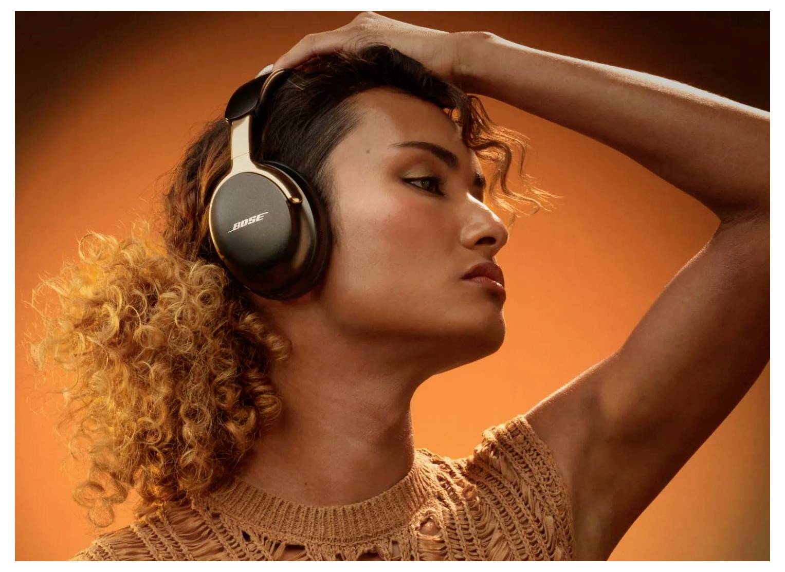 Woman wearing Bose headphones against an orange background, posing with one arm raised over her head.