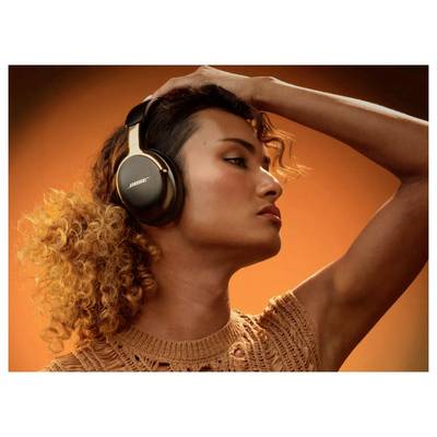 Woman wearing Bose headphones against an orange background, posing with one arm raised over her head.