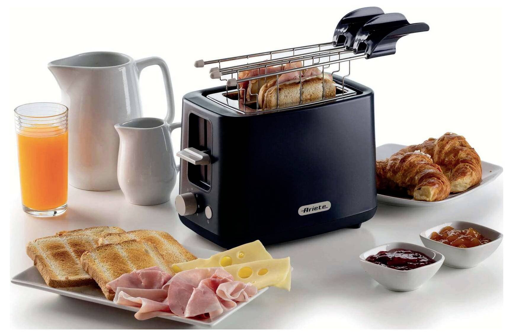Breakfast spread with a toaster toasting bread, croissants, orange juice, ham, cheese, jam, and small pitchers on a white background.