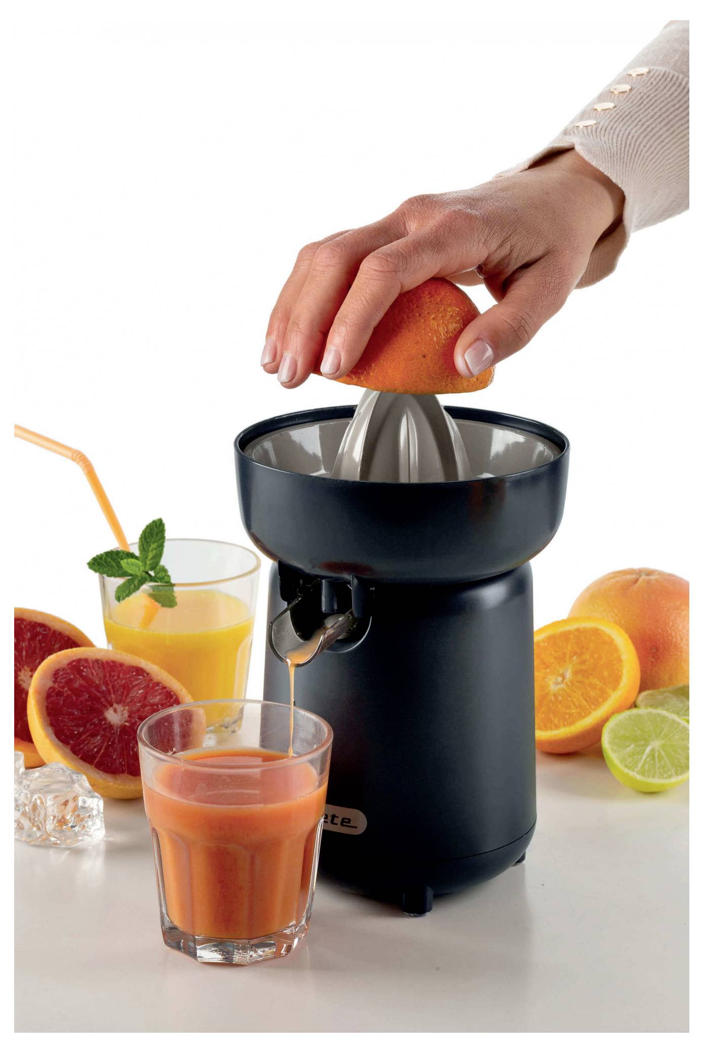Hand squeezing an orange on a black juicer, with fresh citrus slices and two glasses of juice nearby.