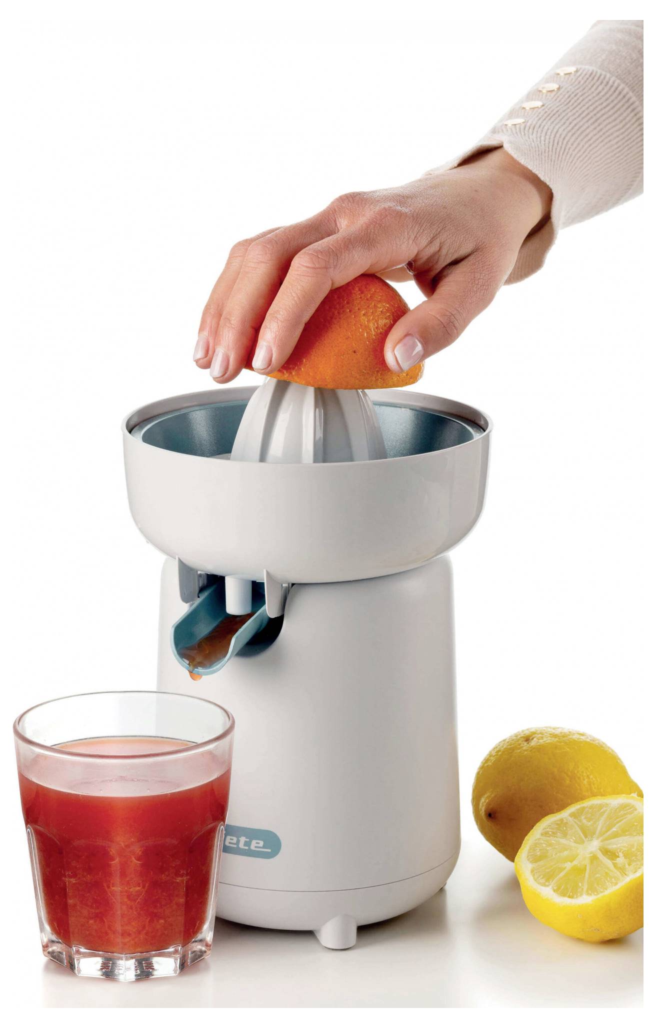Hand squeezing an orange on a juicer, with a glass of red juice and a cut lemon beside it.