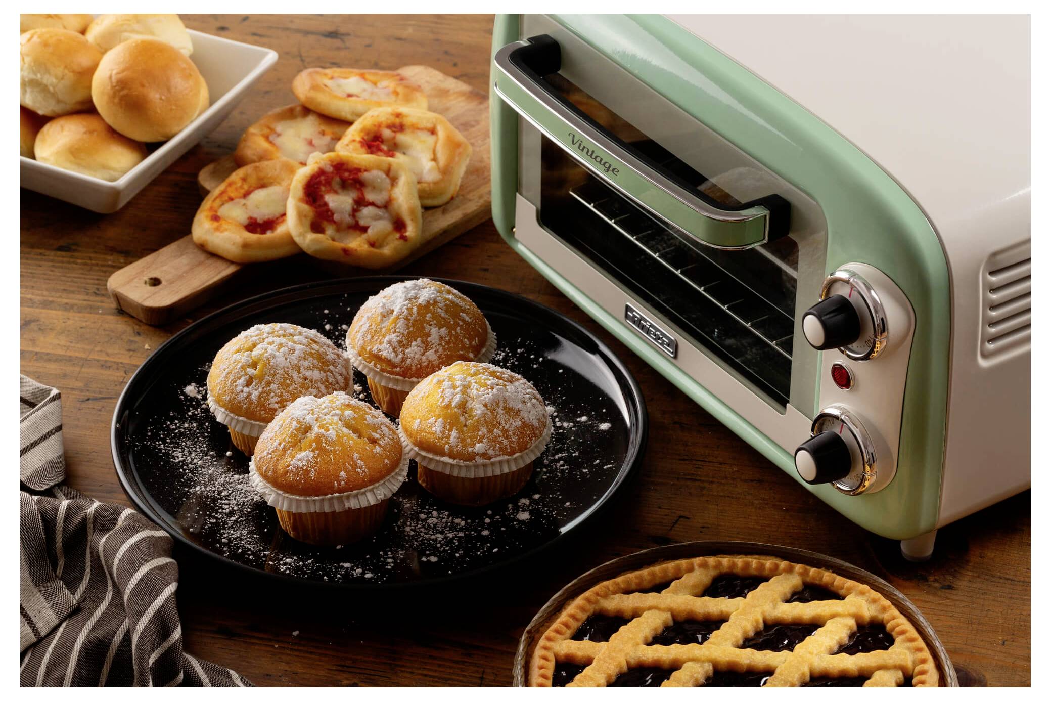 Vintage mint-green toaster oven beside a plate of powdered muffins and assorted baked pastries on a wooden table.<br>