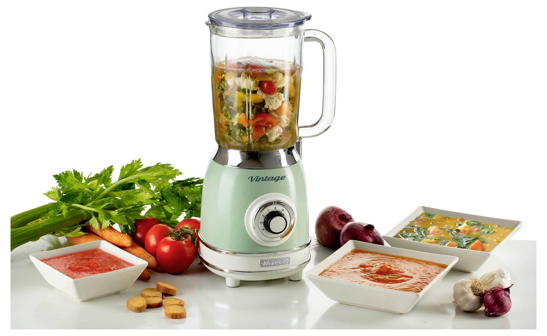 Mint-green blender with chopped vegetables inside, surrounded by bowls of soup ingredients, fresh herbs, tomatoes, croutons, and garlic on a white surface.