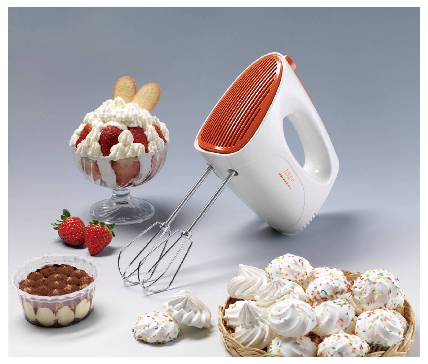 Electric hand mixer with beaters beside assorted desserts, including strawberry parfait, cakes, meringues, and a basket of frosted treats.